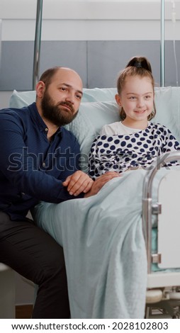 Hospitalized sick little patient lying in bed with family watching entertainment movie on television during medical recovery examination in hospital ward. Kid waiting for disease treatment