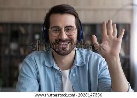 Sincere smiling young businessman in glasses and wireless headphones looking at camera, waving hand making hello gesture starting online video call conversation, communicating distantly with colleague Royalty-Free Stock Photo #2028147356