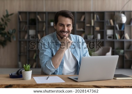 Portrait of smiling young successful male freelancer posing in modern office, sitting at table with computer. Happy millennial businessman looking at camera, feeling excited of new challenges.