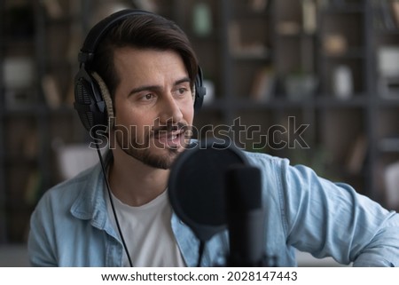 Creative young man wearing wired headphones, singing own song in professional stand microphone. Happy millennial male musician performing recording audio or practicing vocal, hobby activity concept.