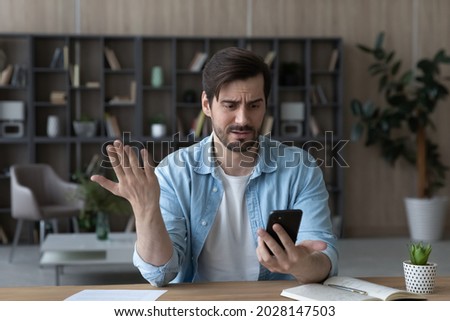 Unhappy young man looking at phone, feeling nervous of bad device work, internet disconnection, lost data or inappropriate online content. Anxious male user dissatisfied with application or service. Royalty-Free Stock Photo #2028147503