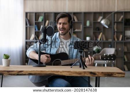 Happy young inspired man playing guitar, singing song in professional stand mic, recording own audio performance on smartphone web camera. Professional blogger teacher streaming live video online.