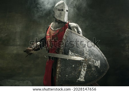 Noble warrior. Portrait of one medeival warrior or knight in armor and helmet with shield and sword posing isolated over dark background. Royalty-Free Stock Photo #2028147656