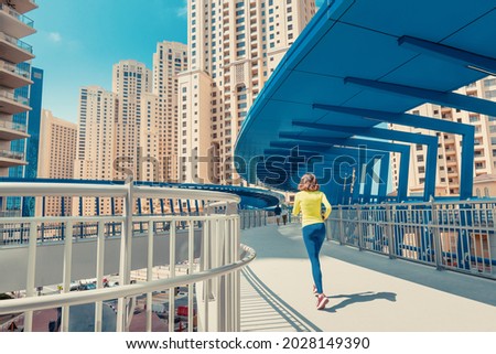 Young woman in bright sportswear quickly runs across a pedestrian bridge in the Dubai Marina district. The concept of a female healthy lifestyle and fitness Royalty-Free Stock Photo #2028149390