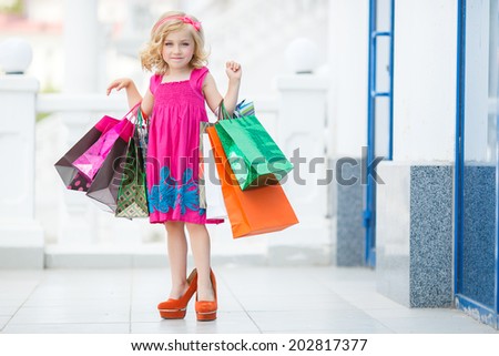 Little fashion girl with packages in a large shopping center. Pretty smiling little girl with shopping bags with thumb up sign in the shop