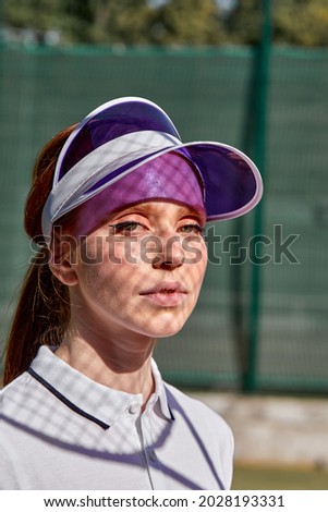 Close-up portrait of pretty young, european woman in cap confidently looking at camera, on tennis court before competition. Determined to win. Dressed in stylish sports tennis uniform white dress.