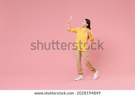 Full length side view of young smiling brunette latin woman 20s in yellow shirt doing selfie shot on mobile phone walk show victory v-sign gesture isolated on pastel pink background studio portrait