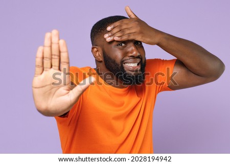 Confused displeased young african american man wearing basic casual orange t-shirt showing stop gesture with palm put hand on head looking camera isolated on pastel violet background studio portrait