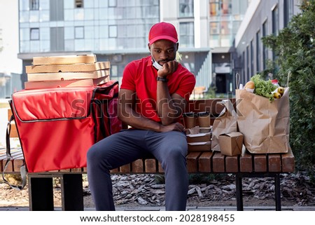 Bored and sad African american young Courier in red uniform with thermal backpack delivering fresh lunches to clients, sit on bench waiting outdoors. Takeaway restaurant food delivery concept.