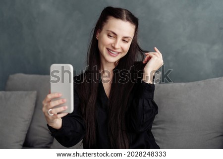 Beautiful european smiling young woman feeling gorgeous while making selfie on modern smartphone while sitting on grey sofa at home, wearing casual clothes. Blogging and modern technologies concept