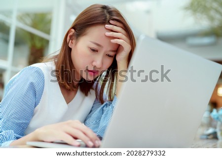 A beautiful Asian woman in a white shirt sits in front of a laptop computer with a stressed face clutching her head while working in a bakery.