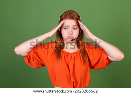 Young red hair woman in casual orange blouse on green background manager call centre help line worker with headphones talk tired exhausted