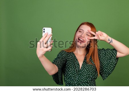 Young red hair woman on green background with mobile phone take photo selfie photographing herself  having fun
