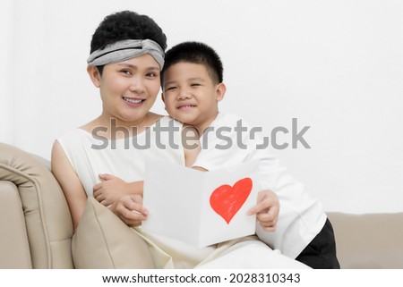 Cute Asian little boy son congratulating his mom happy with Mothers day, giving her handmade greeting postcard with red heart while sitting together on sofa at home. Family holidays concept