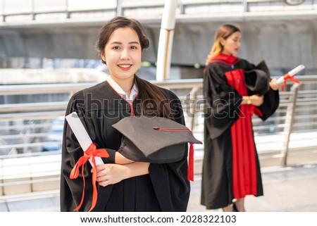 Asian woman in a graduate gown is smiling at the camera while crossing her arm with a certificate in one hand
