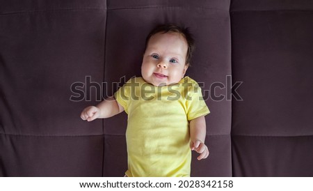 Curious baby girl wearing yellow bodysuit with funny picture lies on back smiling on large brown sofa in spacious apartment room closeup