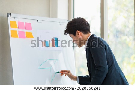 Asian young male professional employee officer in formal suit stand drawing picture on white board presenting analysis data information with graph and chart paperwork with post it in company meeting.