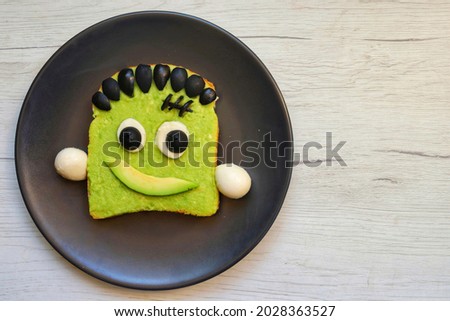 Frankenstein avocado toast made from toast,mashed avocado,mozzarella balls and black olives on black plate with white wood table background.Art food idea for kids party.Top view.Copy space