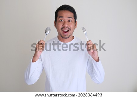 Excited Asian man holding spoon and fork in his hands Royalty-Free Stock Photo #2028364493