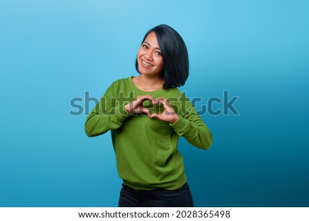 Portrait of smiling Asian woman showing heart gesture with two hands on blue background