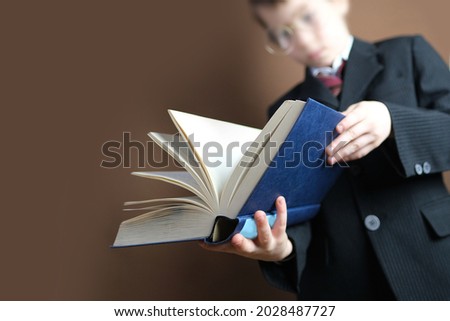 smart child 7-8 years old, kid in black school suit stands and reads thick tome, open paper book, selective focus, concept young child prodigy, back to school, gaining knowledge, education