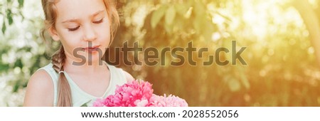 portrait of a happy cute little caucasian seven year old kid girl, holds in hands and love to see a bouquet of pink peony flowers in full bloom on the background of nature. banner. flare