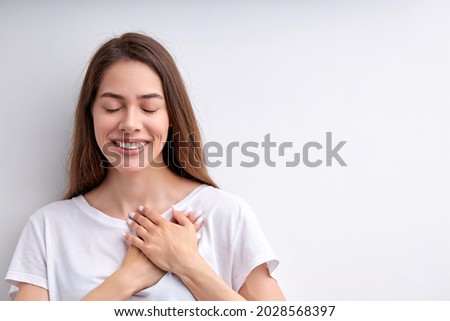 Pretty happy woman expressing gratitude, holding hands on chest, thankful for everything, with eyes closed, having nice smile, casually dressed. isolated on white studio background, copy space Royalty-Free Stock Photo #2028568397