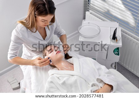 Young pretty woman looking at camera while laying on couch being treated by cosmetologist removing cosmetology mask on face, side view portrait. Professional confident caucasian beautician at work