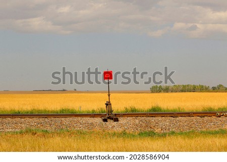 Landscape image of a red railroad track signal device next to the railroad tracks with a gold field behind it.