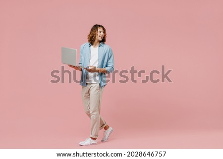 Full length young minded happy man with long curly hair wear blue shirt white t-shirt hold use work on laptop pc computer look aside isolated on pastel plain pink color wall background studio portrait