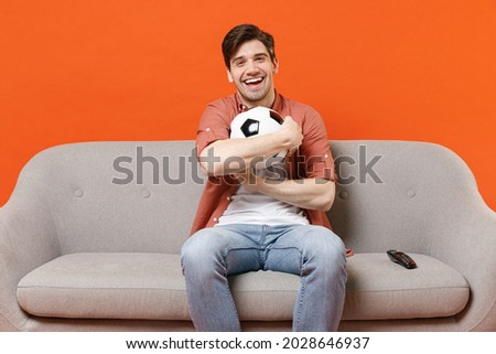 Young smiling man football fan in shirt support team sit on sofa home watching tv live stream hold hug soccer ball isolated on orange background studio portrait People lifestyle sport leisure concept