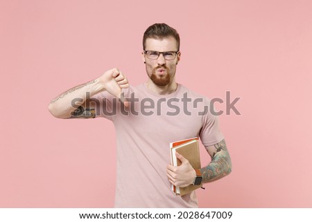Displeased young bearded tattooed man guy in pastel casual t-shirt, eyeglasses isolated on pink background studio portrait. People lifestyle concept. Mock up copy space. Hold books showing thumb down