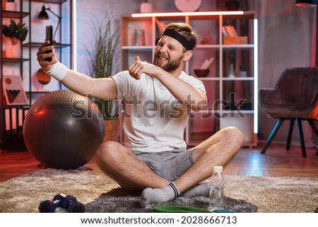 Likable smiling relaxed young bearded sportsman in training clothes sitting with crossed legs on the floor at home and making funny selfies, grimacing on his smartphone