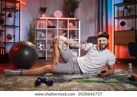Healthy lifestyle concept. Handsome happy smiling sporty young bearded Caucasian man in sportswear lying on the floor in the evening and ready to do exercises.