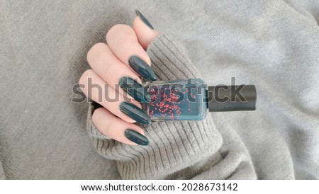Female hand with long nails and dark blue teal manicure holds a bottle of nail polish