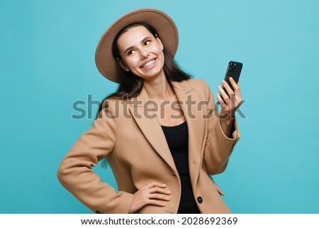 Charming brunette woman in beige classic coat and hat. Lady with smartphone makes selfie on blue background. Blogger influencer looking at camera make video conference call recordin.