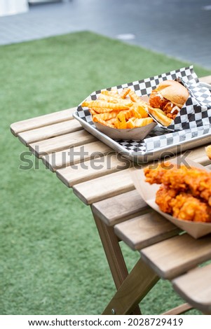A view of a common American street food sitting on a wooden patio table, in an outside area, featuring chicken tenders, crinkle cut french fries, and a chicken burger.
