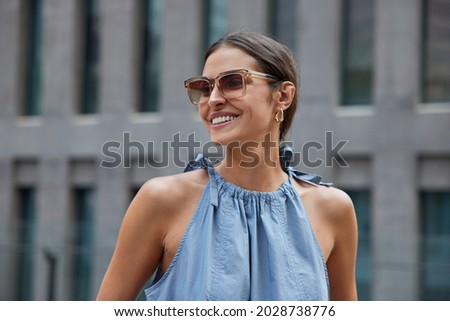 Pleased European woman with dark hair smiles broadly wears sunglasses blue dress enjoys summer walk in urban setting glad to meet friend outside expresses positive emotions returns glad after shopping Royalty-Free Stock Photo #2028738776