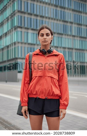 Vertical shot of attractive brunette woman looks seriously at camera walks to gym to have workout carries rolled caremat on shoulder dressed in anorak and shorts poses at street in big city.