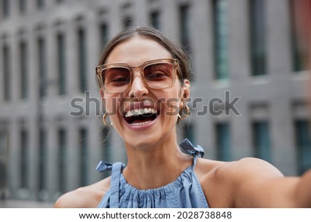 Overjoyed female model laughs happily enjoys spare time takes selfie portrait outside against blurred building background wears sunglasses has excursion in ancient city during summer holidays