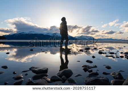 Silhouette of a man standing on a rocky lakeshore in northern Canada with reflection in calm lake below, dramatic clouds and blue sky.  Royalty-Free Stock Photo #2028755117