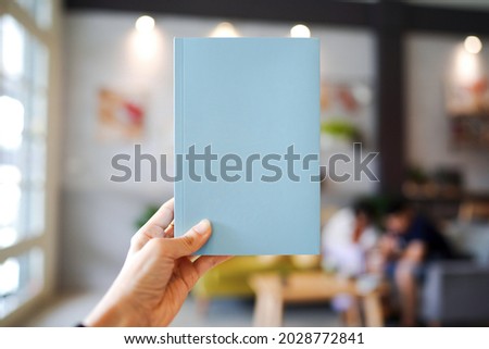 Woman hand holding a light blue book with blank cover for inserting text on blurred cafe background.