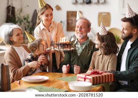 Happy big caucasian family wearing party hats celebrating grandfathers birthday indoors, happy elderly man grandpa holding cake with lit candles, receiving congratulationg during celebration at home