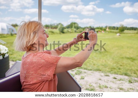 Happy senior woman taking photos via smartphone while riding in open touristic car at natural park during summer tour, active retirement, fulfillment in every age, solo traveler, summer lifestyle