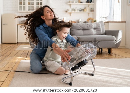Cheerful young mom or nanny have fun with preschool boy at home sitting together in front of big fan dlowing cooling wind in living room. Carefree caring woman of 30 spend time with little son bonding Royalty-Free Stock Photo #2028831728