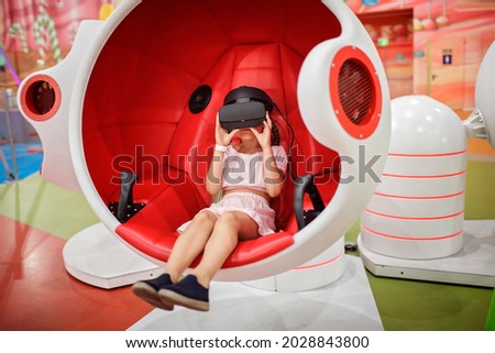 Young girl wearing virtual reality googles and experiencing virtual reality in moving interactive chair at amusement park, future today, new entertainment, indoor lifestyle Royalty-Free Stock Photo #2028843800