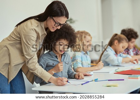Helpful young female teacher showing how to do task to little schoolboy. Kids sitting at the table, studying in elementary school classroom Royalty-Free Stock Photo #2028855284