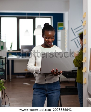 Focused designer standing in digital creative agency office holding laptop typing multimedia production project. Videographer creator editor editing digital montage using production software