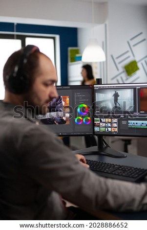 Video maker with headset working with footage and sound on pc with two displays, taking notes while editing customer creative video. Man editor processing film montage in startup agency office.