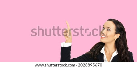 Happy smiling looking up woman in black confident suit, showing pointing advertising at copy space. Business concept. Rose pink color background. Attractive brunette businesswoman. Female bank manager